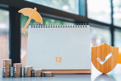 Zdjęcie przedstawiające biały kalendarz z liczbą 12, obok którego znajdują się rosnące stosy monet, mały żółty parasol oraz drewniana tarcza z symbolem „v”. Całość symbolizuje planowanie finansowe, oszczędzanie i ochronę kapitału.
