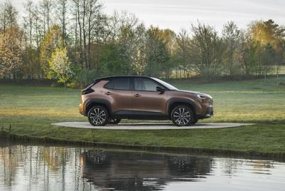 Toyota Yaris Cross w kolorze miedzianym zaparkowana na trawie nad brzegiem wody w otoczeniu zielonych drzew.