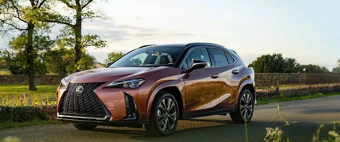 Zdjęcie przedstawia Lexusa UX 300h w kolorze miedzianym, z czarnym dachem i elementami wykończenia. Auto jedzie po asfaltowej drodze, w tle widać zielone pobocza oraz drzewa. 