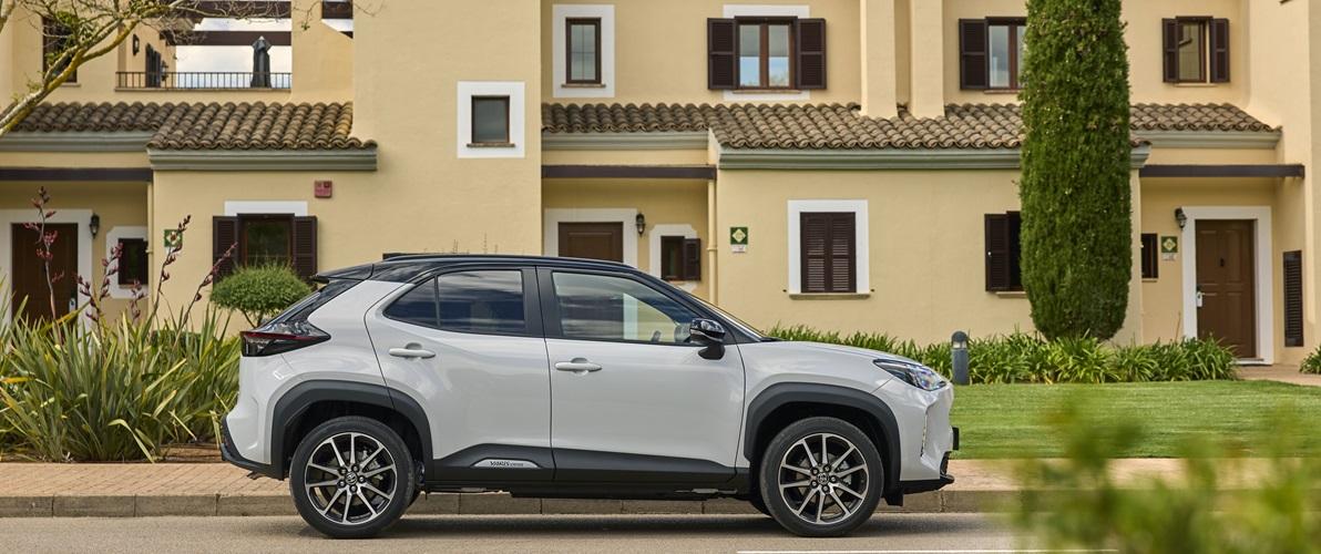 Zdjęcie przedstawia szarą Toyotę Yaris Cross, zaparkowaną przed budynkiem w żółtych kolorach. Budynek składa się z 3 osobnych mieszkań, ma brązowe drzwi oraz okiennice. Pomiędzy budynkiem a autem znajduje się trawnik. Widok na auto z boku.