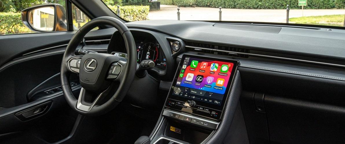 Zdjęcie z wnętrzem samochodu Lexus z widokiem na deskę rozdzielczą i kierownicę. Na środku deski rozdzielczej znajduje się ekran dotykowy, na którym wyświetlane są ikony aplikacji. Kierownica jest wielofunkcyjna, z logo Lexusa pośrodku.