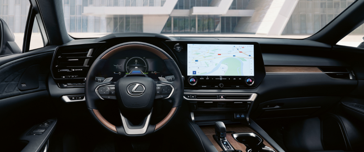 Wnętrze samochodu Lexus RX, z widocznym kokpitem, kierownicą, deską rozdzielczą i ekranem nawigacji. Wnętrze jest eleganckie i nowoczesne, z elementami wykończenia z drewna i skóry. Na ekranie nawigacji widoczna jest mapa z zaznaczoną trasą.