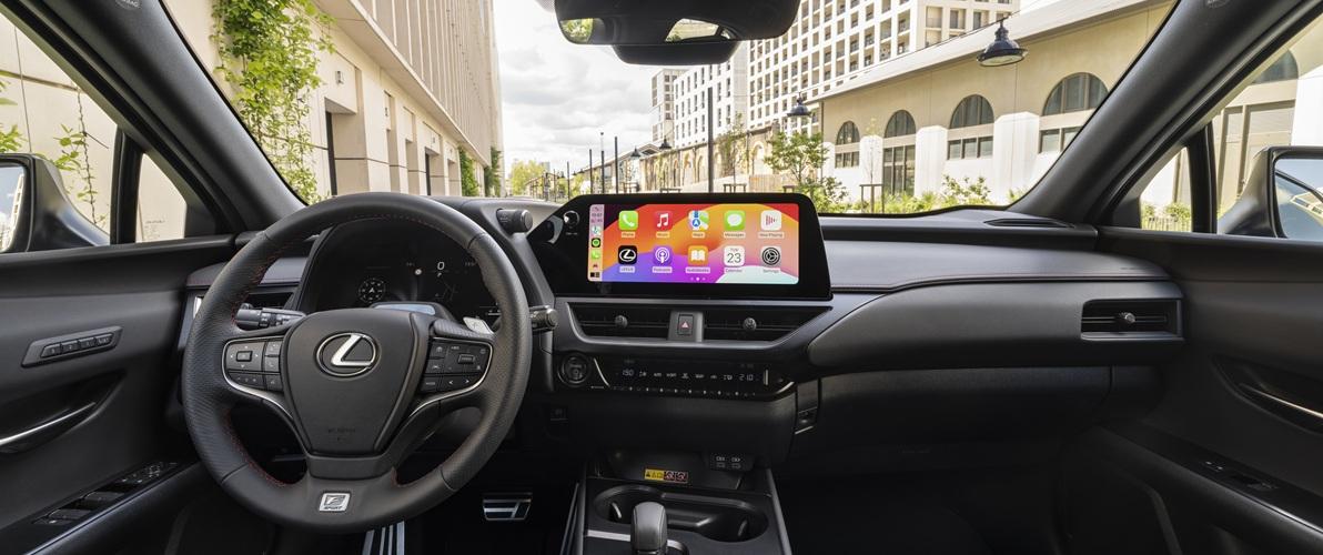 Zdjęcie wnętrza samochodu marki Lexus, z widocznym kokpitem i deską rozdzielczą. Na centralnym ekranie widoczny jest interfejs Apple CarPlay z ikonami aplikacji. Kierownica ma logo Lexusa i oznaczenie "F SPORT". Za szybą przednią widać fragment ulicy z budynkami.