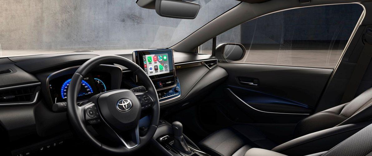 Wnętrze samochodu Toyota Corolla, widok z miejsca kierowcy. Widać kierownicę z logo Toyoty, cyfrowy zestaw wskaźników oraz duży, centralny ekran dotykowy na desce rozdzielczej. Wnętrze jest utrzymane w ciemnej kolorystyce z subtelnym niebieskim oświetleniem ambientowym na panelach drzwi.