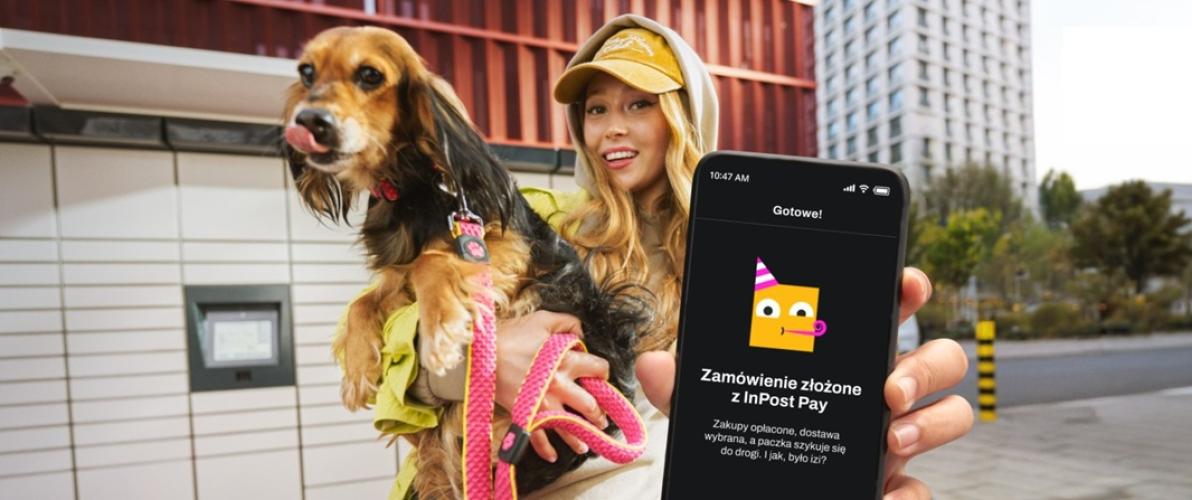 Uśmiechnięta dziewczyna trzymająca dużego psa na jednej ręce, w drugiej pokazująca smartfona z ikoną inpost. W tle widać paczkomat. 