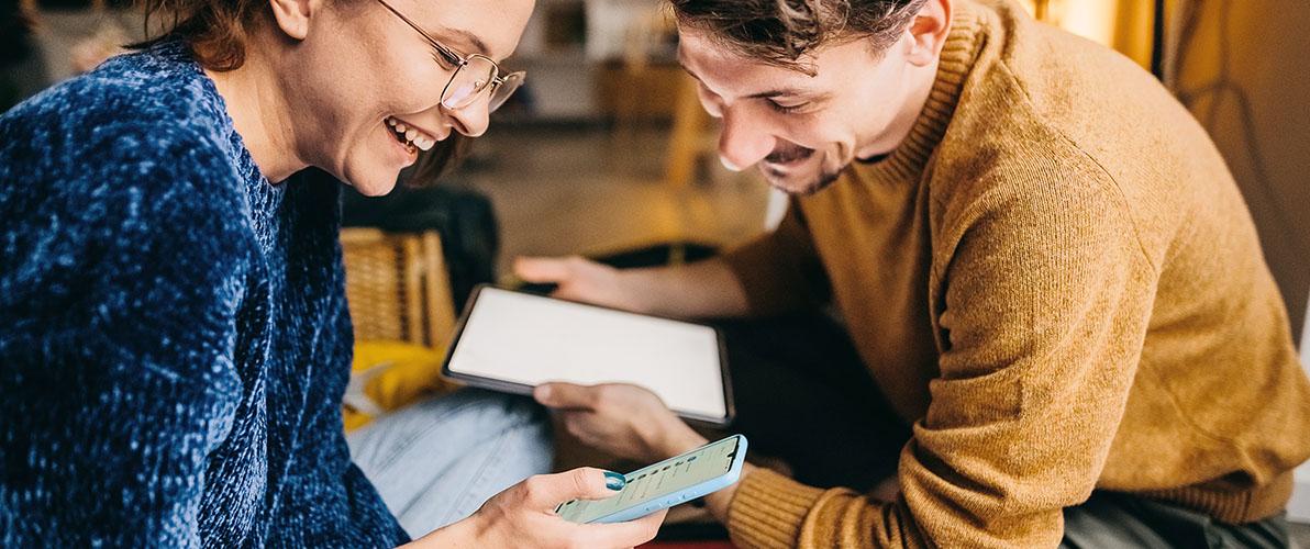 Uśmiechnięta kobieta w okularach i niebieskim swetrze oraz mężczyzna w żółtym swetrze wspólnie przeglądają zawartość tabletu i smartfona.