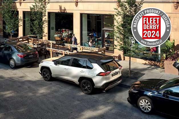 Trzy samochody zaparkowane jeden za drugim na ulicy, pośrodku biała Toyota RAV 4. Obok znajduje się budynek oraz drzewa