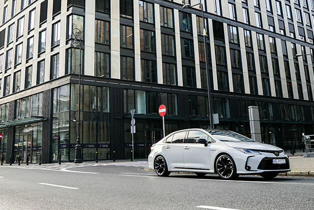 Biała Toyota Corolla Sedan, zaparkowana na ulicy przed nowoczesnym budynkiem biurowym. Budynek ma ciemne okna i jasne elementy konstrukcyjne. Na ulicy widać oznakowanie poziome oraz znak drogowy "zakaz wjazdu"