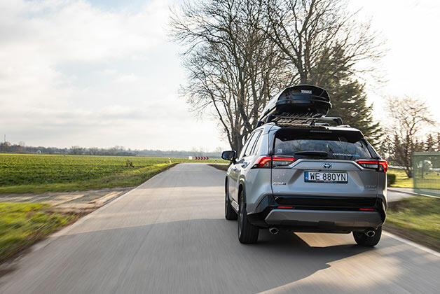 Srebrna Toyota RAV4, jadąca po asfaltowej drodze. Na dachu widoczny jest dodatkowy bagażnik. Na poboczach widoczne zielone łąki, po prawej stronie rosnących kilka wysokich drzew.