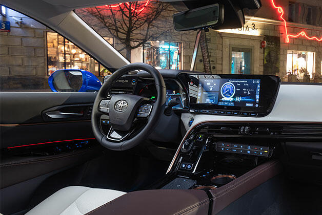 Wnętrze Toyoty. Widoczna jest deska rozdzielcza z kierownicą, ekranem informacyjno-rozrywkowym oraz elementami sterowania. Wnętrze utrzymane jest w tonacji brązowo-białej z elementami czerni i srebrnymi akcentami.