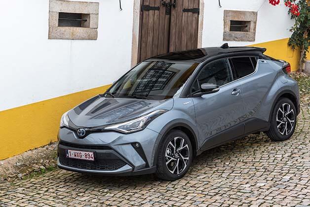 Na zdjęciu znajduje się srebrny samochód Toyota C-HR, zaparkowany na brukowanej nawierzchni przed budynkiem o biało - żółtej ścianie i z drewnianymi drzwiami.
