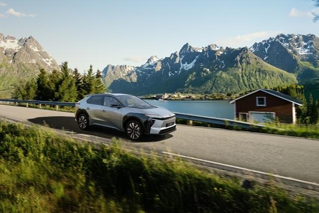 Na zdjęciu widoczny jest srebrny samochód Toyota bZ4X, elektryczny SUV, który jedzie po asfalcie. W tle rozciąga się malowniczy krajobraz z górami i jeziorem. Tuż za drogą widać mały, drewniany domek w kolorze brązowym z białymi wykończeniami.