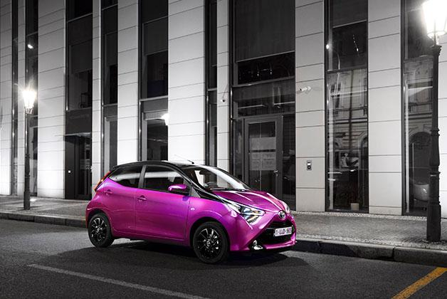 Zdjęcie przedstawia Toyotę Aygo w kolorze różowym, zaparkowaną na ulicy w nocy. Samochód ma czarne felgi i reflektory LED. W tle widoczny jest budynek z dużymi oknami i latarnie uliczne.
