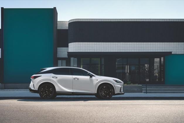 Biały samochód Lexus RX, zaparkowany na ulicy przed nowoczesnym budynkiem, z elementami z ułożonych białych kafelków oraz turkusowymi ścianami. Widoczne szklane wejście.