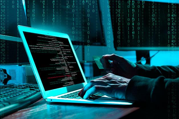 Ręce osoby pracującej przy laptopie, na ekranie kod komputerowy, w tle monitory – jedna dłoń trzyma kartę płatniczą; ilustracja zagrożeń związanych z bezpieczeństwem danych.