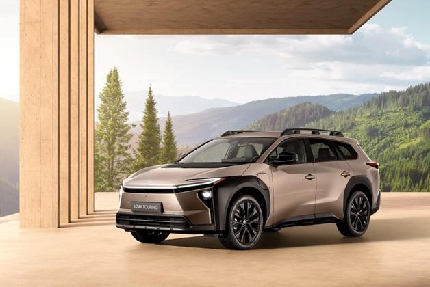 Na zdjęciu widoczny jest elektryczny samochód Toyota bZ4X touring z 2025 roku. Auto ustawione jest pod ukosem, z widocznym przodem i lewym bokiem. W tle widoczne zielone drzewa i góry. Zdjęcie zrobione jest w słoneczny dzień.