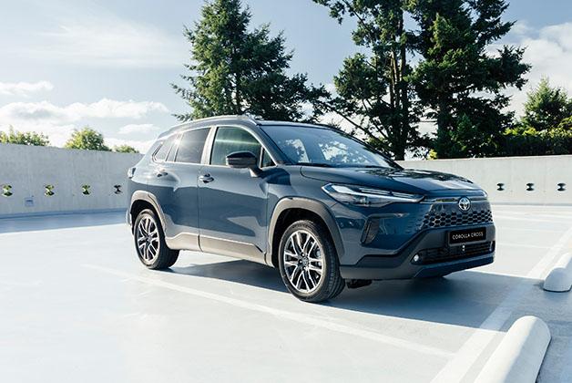  Szara Toyota Corolla Cross zaparkowana na górnym piętrze parkingu. W tle widać drzewa, a w prawym dolnym rogu betonowy ogranicznik miejsca parkingowego. Zdjęcie zostało zrobione w słoneczny dzień.