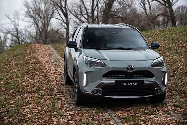 Toyota Yaris Cross, w kolorze srebrnym, sfotografowana w jesiennym otoczeniu. Samochód stoi na lekko pagórkowatej drodze, która jest obficie pokryta opadłymi, suchymi liśćmi.