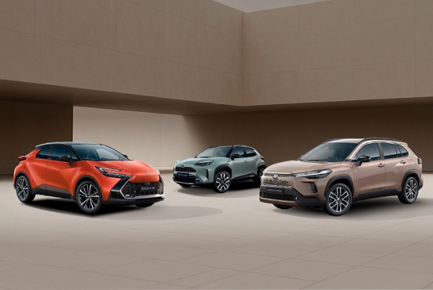 Trzy crossovery Toyoty są zaparkowane w pomieszczeniu. Od lewej strony widoczny jest Toyota C-HR w kolorze czerwonym, pośrodku Toyota Yaris Cross w kolorze szarym, a po prawej stronie Toyota Corolla Cross w kolorze beżowym. Wszystkie pojazdy mają włączone światła. Tłem jest minimalistyczne wnętrze z beżowymi i brązowymi ścianami.