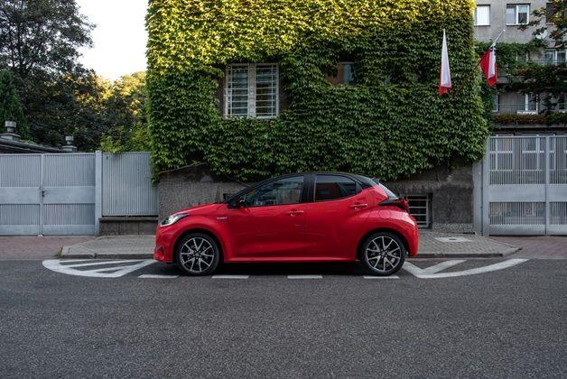 Czerwony hatchback - Toyota Yaris - zaparkowany na ulicy, na tle budynku całkowicie pokrytego bluszczem, z widoczną polską flagą.