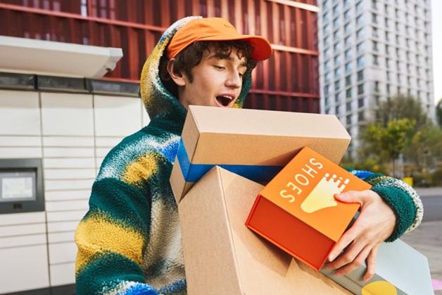 Młody, uśmiechnięty mężczyzna, trzyma w ramionach kilka pudełek po butach, idąc ulicą w mieście. Na jednym z pudełek widnieje napis "SHOES" oraz grafika stopy. W tle widać paczkomat.