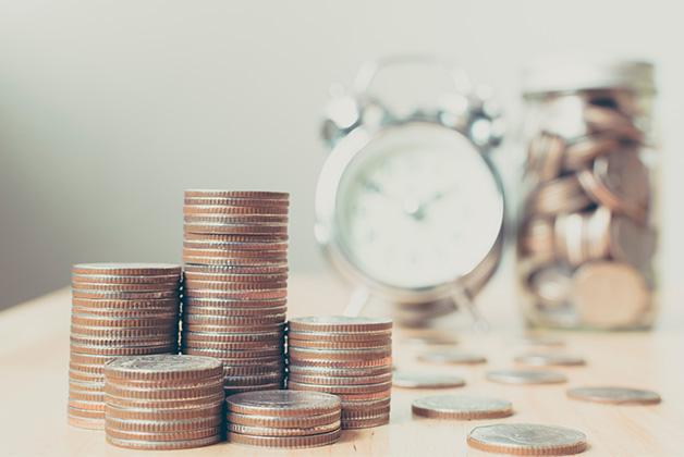 Zdjęcie przedstawia stosy monet, budzik i słoik z pieniędzmi na drewnianym stole, ilustrujące koncepcję oszczędzania oraz lokat terminowych.
