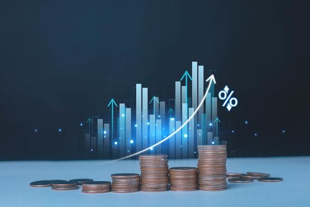 Ułożone monety w rosnących słupkach symbolizujące zysk lub oszczędności, przed którymi znajduje się cyfrowy, rosnący wykres słupkowy i strzałka w górę z symbolem procentowym.