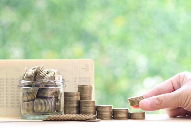 Ręka umieszczająca mały stos złotych monet na szczycie rosnącego rzędu stosów monet, z słoikiem pełnym monet i otwartym zeszytem oszczędnościowym w tle. W tle zielone rozmyte liście.