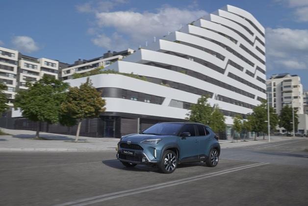 Niebieski samochód Toyota Yaris Cross jedzie ulicą miejską w słoneczny dzień. W tle znajduje się nowoczesny, biały budynek mieszkalny o falistej architekturze oraz inne bloki mieszkalne i drzewa.