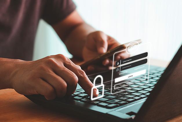 Zdjęcie przedstawia ręce osoby wpisującej dane na klawiaturze laptopa. Obok leży smartfon z ekranem logowania i symbolem kłódki, co sugeruje bezpieczeństwo online.
