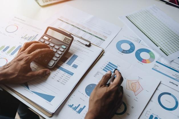 Ręce biznesmena lub księgowego używającego kalkulatora i długopisu do obliczania finansów, pracującego nad dokumentami z wykresami i danymi.