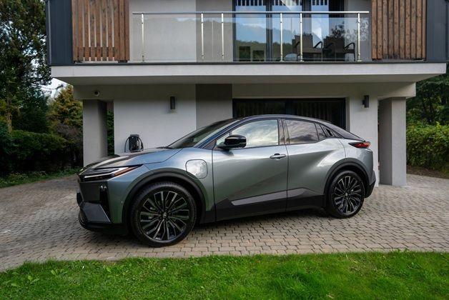 Zdjęcie przedstawia srebrnoszary, w pełni elektryczny samochód Toyota C-HR+ SUV, zaparkowany na podjeździe przed nowoczesnym domem. Pojazd ma futurystyczny design.