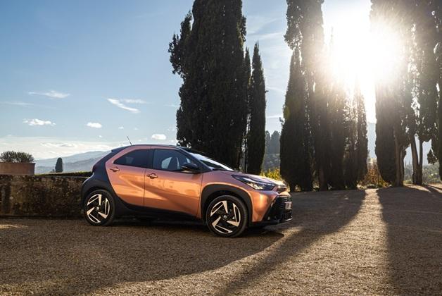 Pomarańczowo-czarna Toyota Aygo X zaparkowana na żwirowym podjeździe, w otoczeniu wysokich cyprysów, pomiędzy którymi świeci słońce.