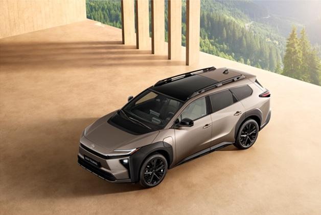 Widok z góry na elektryczny samochód Toyota bZ4X Touring (SUV lub kombi) w kolorze brązowym lub szampańskim, zaparkowany na beżowym, nowoczesnym dziedzińcu. Samochód ma czarny dach, relingi dachowe, czarne nadkola i felgi, a w tle widać górzysty, zalesiony krajobraz.