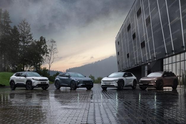 Cztery elektryczne SUV-y Toyota bZ4X Touring w różnych kolorach (od lewej: biały, szary, biały i brązowy) zaparkowane w rzędzie na mokrej, wybrukowanej powierzchni obok nowoczesnego budynku, z zielonymi wzgórzami i zachmurzonym niebem w tle.