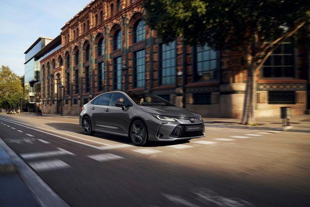 Zdjęcie przedstawia ciemnoszary samochód Toyota Corolla Sedan jadący ulicą obok dużego, starego budynku z cegły z wieloma oknami. W aucie widoczne są dwie osoby.