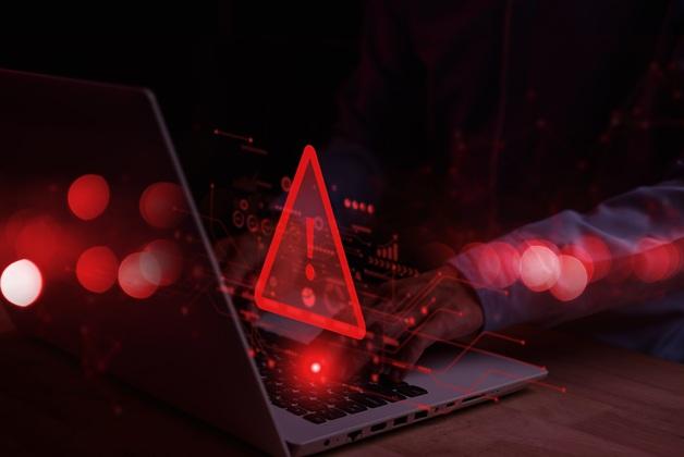 Zbliżenie dłoni osoby pracującej na laptopie w ciemnym otoczeniu, z nałożonym świecącym czerwonym trójkątem ostrzegawczym (ikona zagrożenia cybernetycznego) i rozmytymi czerwonymi światłami w tle, ilustrujące naruszenie bezpieczeństwa.