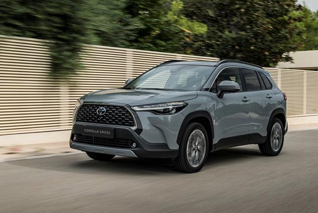 Jasnoszara Toyota Corolla Cross jadąca po asfaltowej drodze na tle nowoczesnego, jasnego ogrodzenia i zielonych drzew.