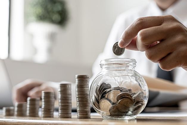 Zdjęcie przedstawia osobę oszczędzającą pieniądze, wrzucającą monetę do szklanego słoika, obok rosnących stosów monet.