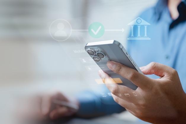 Zdjęcie przedstawia osobę korzystającą ze smartfona do obsługi bankowości mobilnej lub wykonywania płatności online. Na obrazku widoczne są nałożone ikony graficzne symbolizujące proces finansowy: profil użytkownika połączony z budynkiem banku za pomocą linii z „ptaszkiem” (wyrażającym zatwierdzenie lub weryfikację)