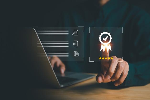Zbliżenie na dłoń osoby przy laptopie, wskazującą na unoszącą się, cyfrową ikonę złotego medalu z białym ptaszkiem (check-mark) i pięcioma gwiazdkami, symbolizującą certyfikację i wysoką jakość