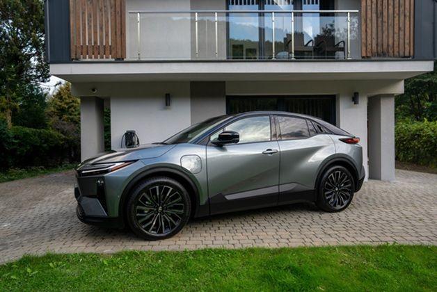 Nowoczesna Toyota C-HR+ w wersji elektrycznej zaparkowana przed budynkiem mieszkalnym z przeszklonym balkonem. Samochód prezentuje dynamiczną sylwetkę typu coupe z charakterystycznymi czarnymi felgami i widocznym gniazdem ładowania na przednim błotniku.