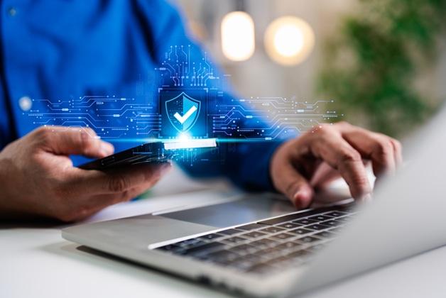 Osoba w niebieskiej koszuli korzystająca z laptopa i smartfona, nad którymi wyświetla się świecąca, niebieska ikona tarczy z haczykiem, symbolizująca bezpieczne połączenie i ochronę danych.