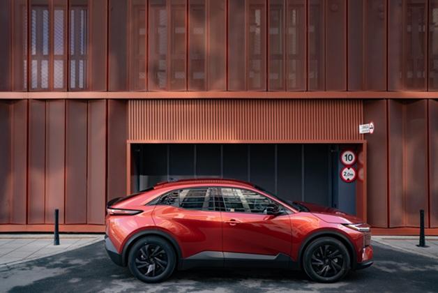 Nowa Toyota C-HR+ w kolorze czerwonym zaparkowana profilem na tle miedzianej, żebrowanej elewacji budynku.