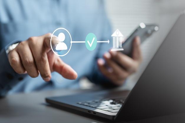 Zdjęcie przedstawia osobę korzystającą z laptopa i smartfona, na które nałożona jest błękitna grafika ilustrująca proces cyfrowej weryfikacji. Ikona użytkownika łączy się poprzez symbol zatwierdzenia (ptaszek w kółku) z ikoną budynku banku, co symbolizuje bezpieczny przepływ danych lub autoryzację transakcji bankowej.