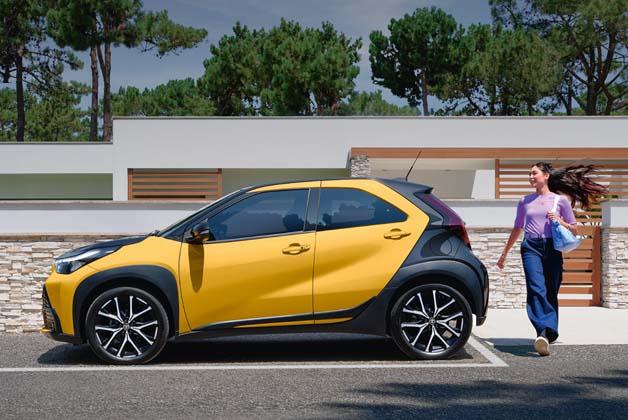 Żółto-czarny miejski crossover Toyota Aygo X zaparkowany profilem na tle nowoczesnego domu. Obok tyłu pojazdu idzie uśmiechnięta kobieta z torbą zakupową, a w tle widać zielone drzewa i błękitne niebo.
