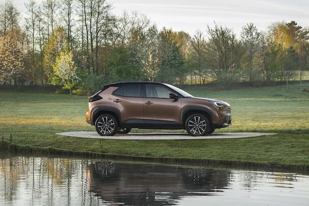Toyota Yaris Cross w kolorze miedzianym zaparkowana na trawie nad brzegiem wody w otoczeniu zielonych drzew.