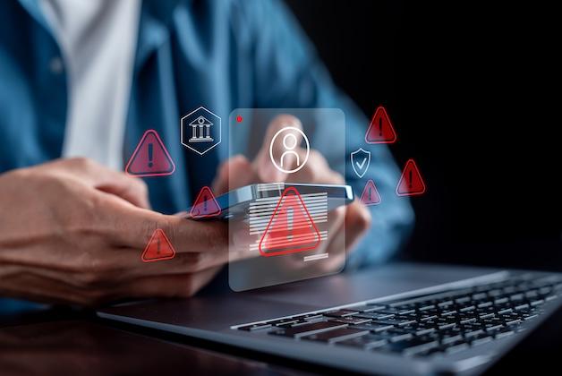 Osoba trzymająca smartfon nad klawiaturą laptopa, otoczona czerwonymi, cyfrowymi ikonami ostrzegawczymi, symbolizująca cyberzagrożenie.
