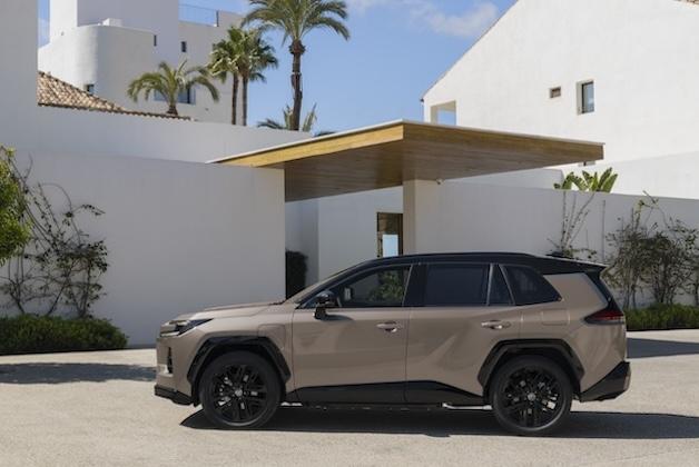Boczny profil nowej beżowo-czarnej Toyoty RAV4 szóstej generacji, zaparkowanej przed nowoczesnym białym budynkiem z drewnianym zadaszeniem w słoneczny dzień