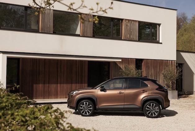 Brązowy SUV Toyota Yaris Cross zaparkowany przed nowoczesnym, białym domem z drewnianą elewacją.